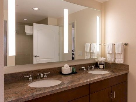 Two sinks on a brown granite countertop, large mirror with vertical lights, towels on rack, and a door in the background.