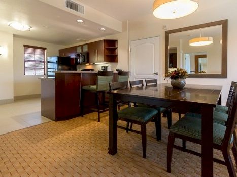 A modern open-concept dining area with a dark wooden table, six green-cushioned chairs, and a contemporary kitchen in the background, breakfast bar included.