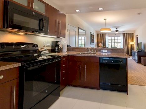 A modern open-concept kitchen with dark wood cabinets, black appliances, a breakfast bar, and a view into a living area with warm lighting.