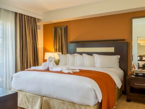 A hotel bedroom with a large bed, white linens, orange accents, a nightstand, lamp, and a balcony view. Top it at 140 characters, always ending the sentence.