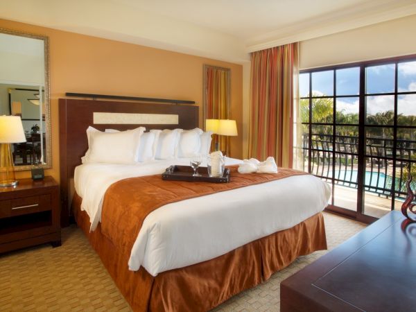 A well-made hotel room with a large bed, brown and white linens, two lamps, a balcony with sliding doors, a dresser, and a mirror.