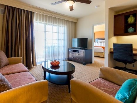 A hotel suite living area with two tan sofas, a round coffee table, a TV on a dresser, a desk nook, large windows with curtains, and a visible bedroom in the background.