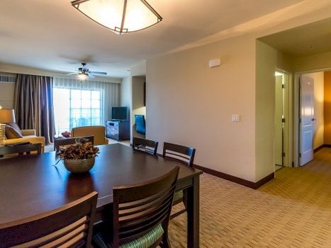 A hotel suite living area with a dark wooden dining table, chairs, a sofa, lamps, a window with curtains, and a doorway to a hallway.