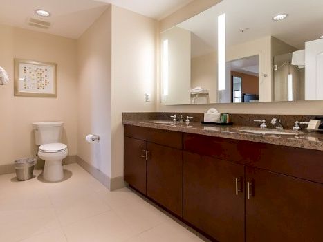 A modern bathroom with a long dark wood vanity, double sinks, large mirror, bright lighting, and a separate toilet area.