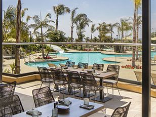 A sunny poolside patio with glass walls, palm trees, lounge chairs, and tables set for dining by the water.