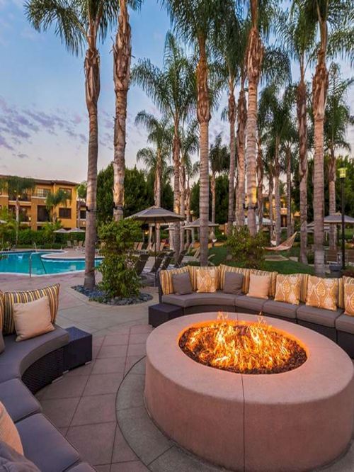 A stylish outdoor lounge with a circular fire pit, curved sectional seating, plush cushions, palm trees, and a swimming pool in the background.
