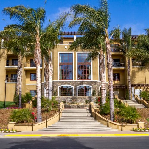 A tropical-style hotel or apartment complex with a grand entrance, palm trees, and warm beige stucco architecture, under a sunny sky.