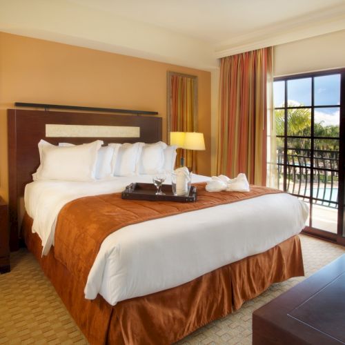 A cozy hotel room with two double beds, warm lighting, a desk, seating area, and large windows with a sunny view.