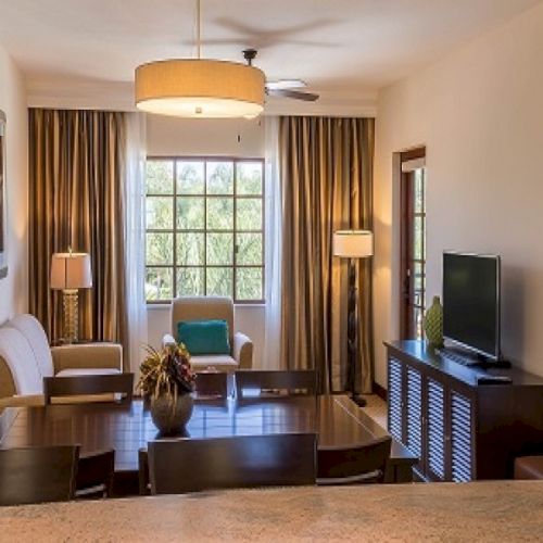 A cozy living room with a sofa, armchairs, coffee table, and a doorway leading to another room, bathed in warm natural light.