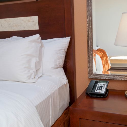 A neatly made hotel bed with white linens, a bedside lamp, and a small wooden nightstand.