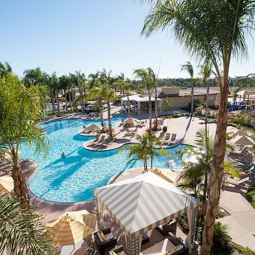 A sunny resort with a blue pool, lounge chairs, palm trees, and a turquoise sea in the background.
